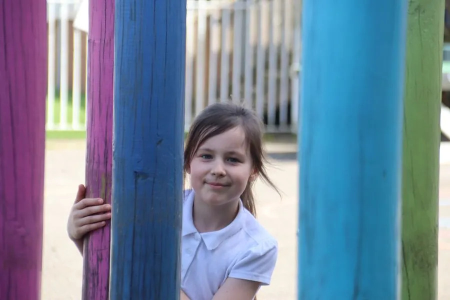 Smiling girl in school uniform peeking out from behind colourful playground posts
