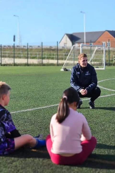 A uSports coach talking to children sitting on the pitch
