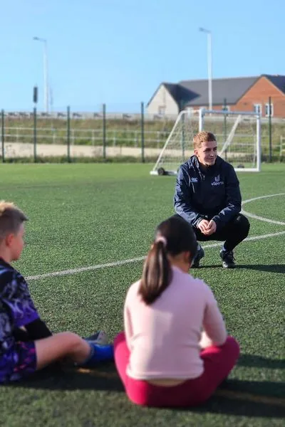 A uSports coach leading a session with children sitting on the pitch